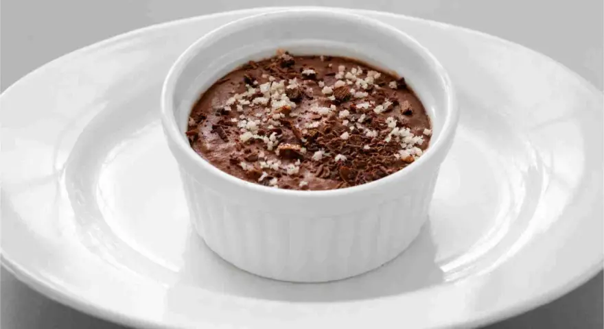 Ein cremiges Mousse au Chocolat, garniert mit Schokoladenraspeln und feinen Salzkristallen, serviert in einer weißen Schale auf einem Teller.