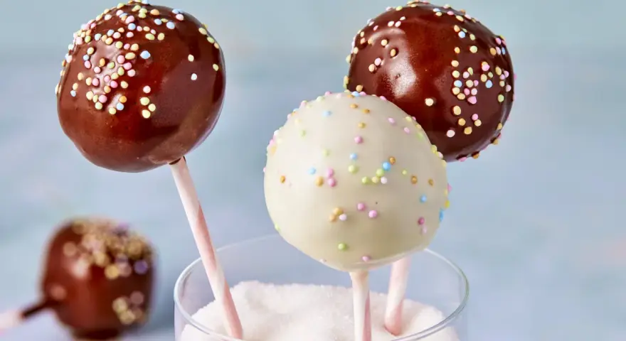Drei bunte cake pops aus kuchenresten, mit Schokolade und Streuseln verziert, stecken in einem Glas voller Zucker.