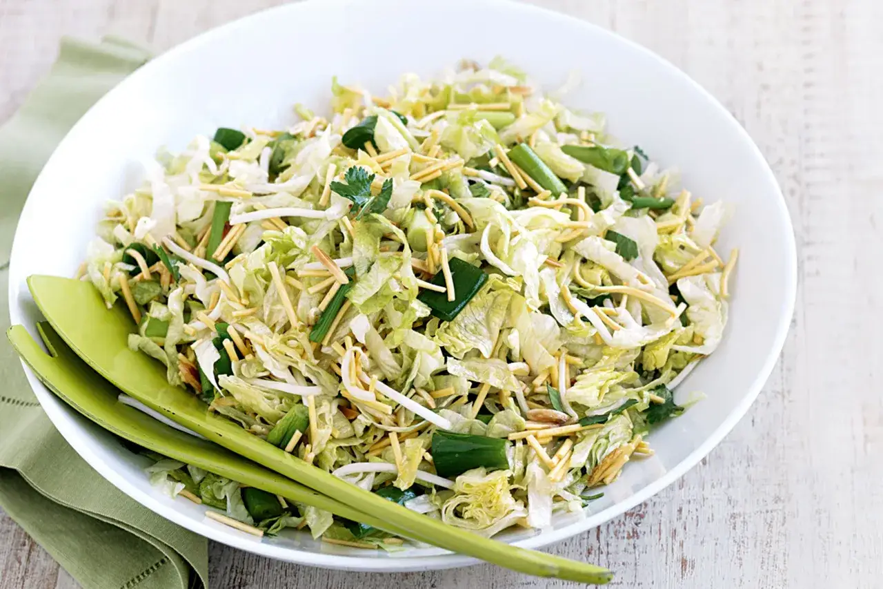 Ein knackiger yum yum Salat mit Eisbergsalat, Sojasprossen und knusprigen Nudeln, serviert in einer weißen Schüssel mit grünen Salatbesteck.