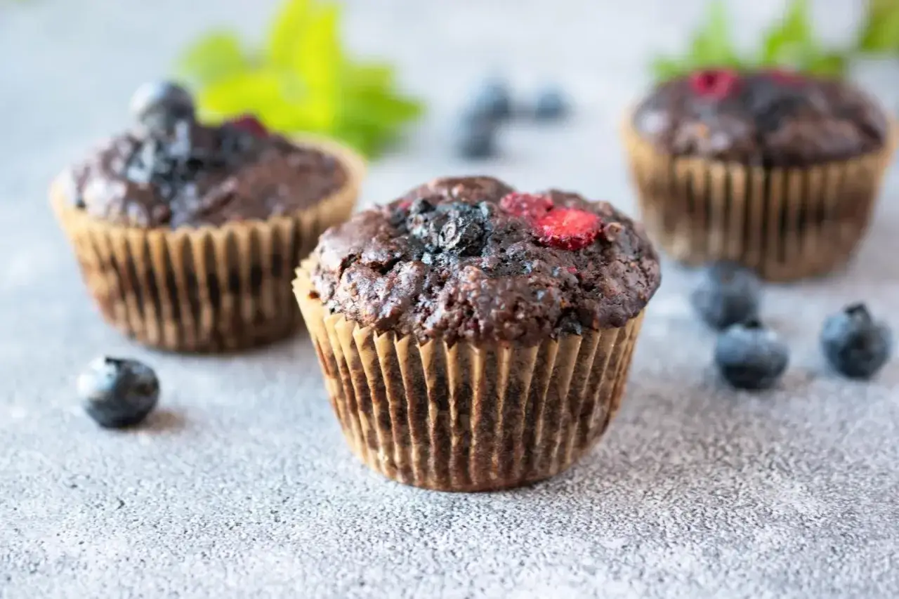 Drei schoko blaubeer muffins mit frischen Beeren und Blaubeeren auf grauem Untergrund.