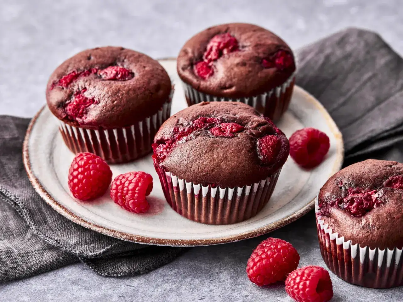 Schoko Himbeer Muffins auf einem Teller, umgeben von frischen Himbeeren und einem grauen Tuch.