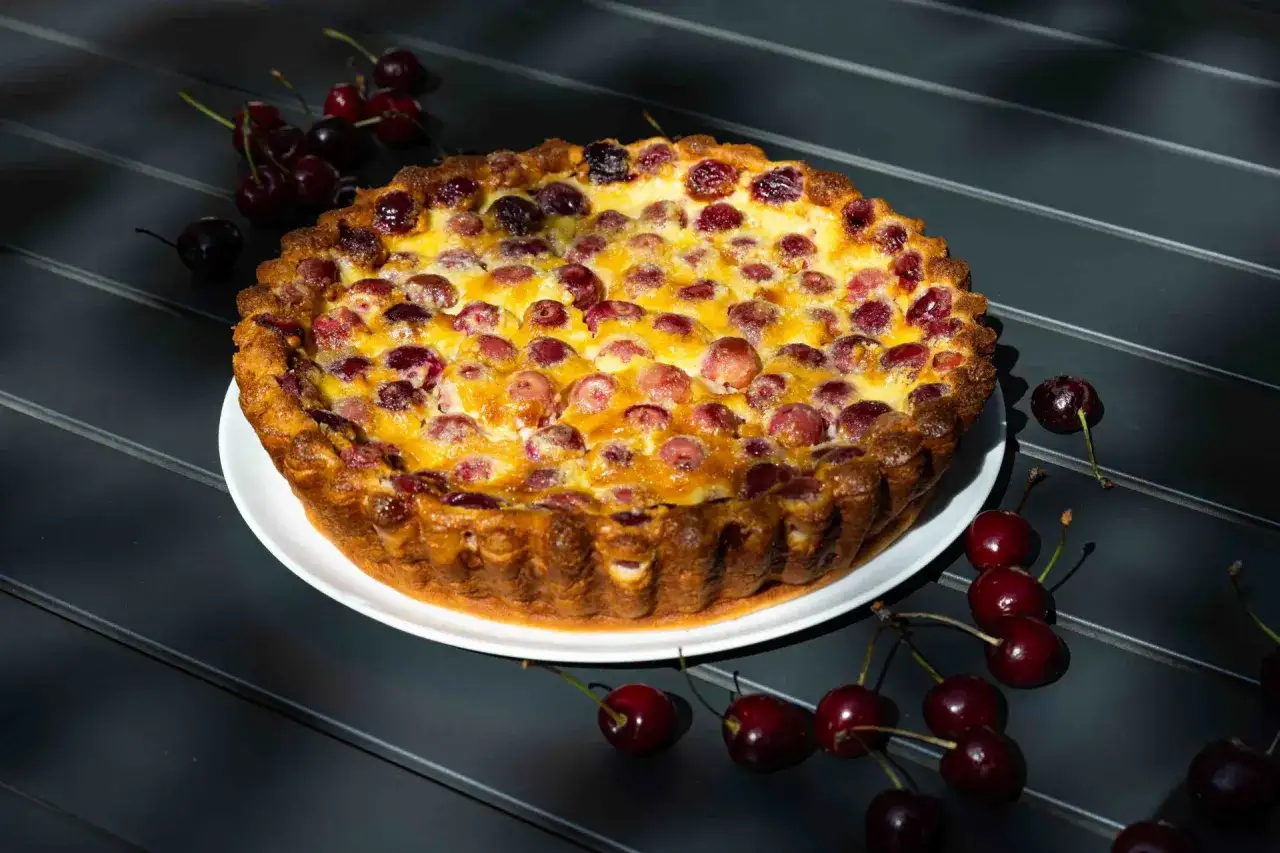 Ein goldbrauner Kirschkuchen, der nach dem clafoutis rezept original duftet, liegt auf einem wei&szlig;en Teller. Frische Kirschen zieren die Oberfl&auml;che und liegen daneben.