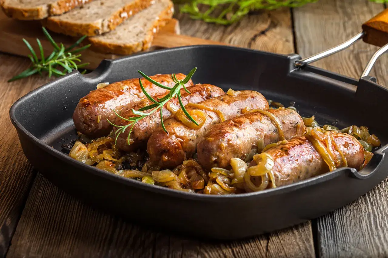 Bratw&uuml;rste in einer Pfanne mit karamellisierten Zwiebeln, wie bei Oma, garniert mit Rosmarin. Dazu Scheiben von dunklem Brot.
