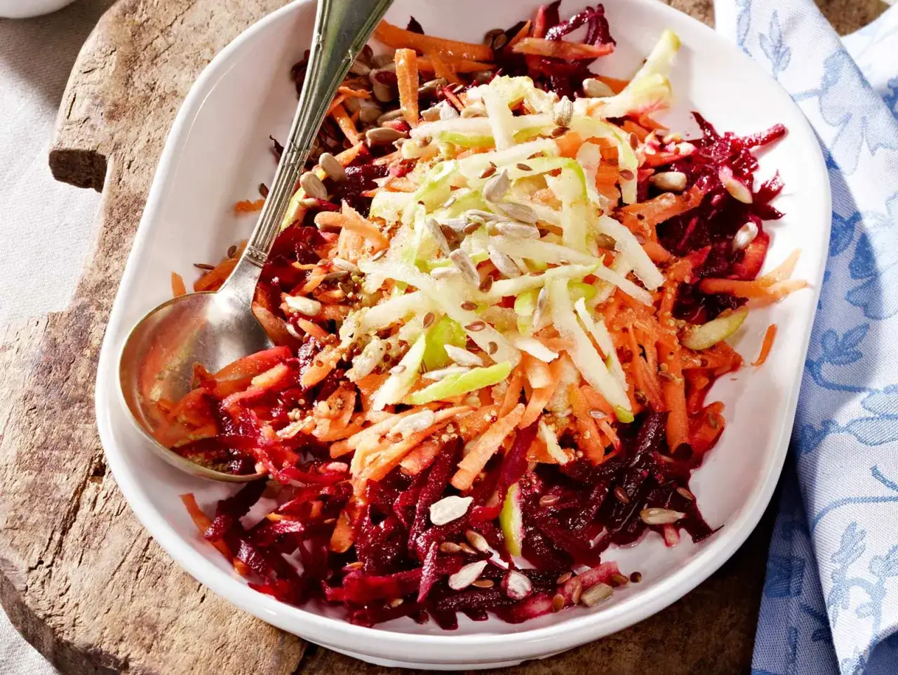Frischer, bunter rote Bete Salat mit Apfel, Karotten und Sonnenblumenkernen in einer wei&szlig;en Sch&uuml;ssel, serviert mit einem L&ouml;ffel.