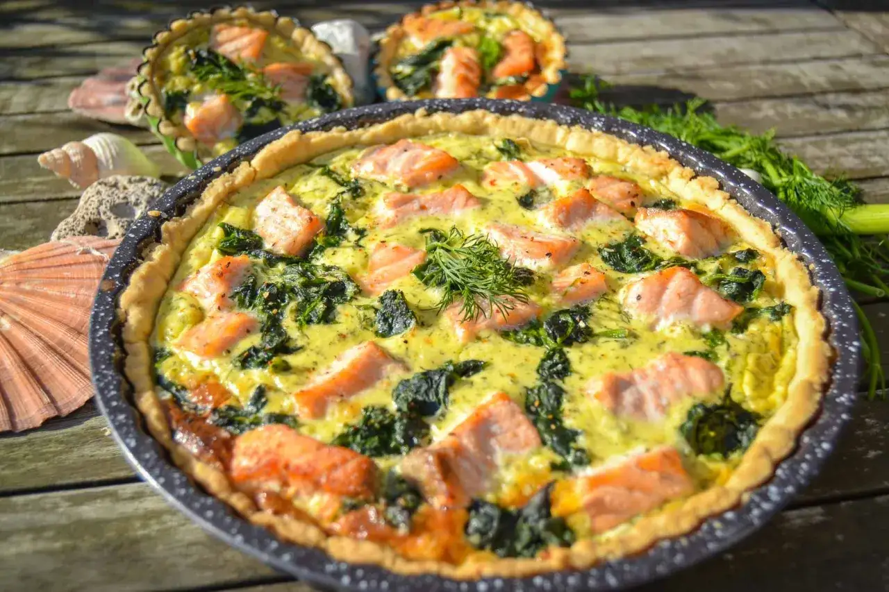 Herzhafte Spinat Lachs Quiche mit Dill garniert, auf einem Holztisch mit Muscheln und Fenchel.