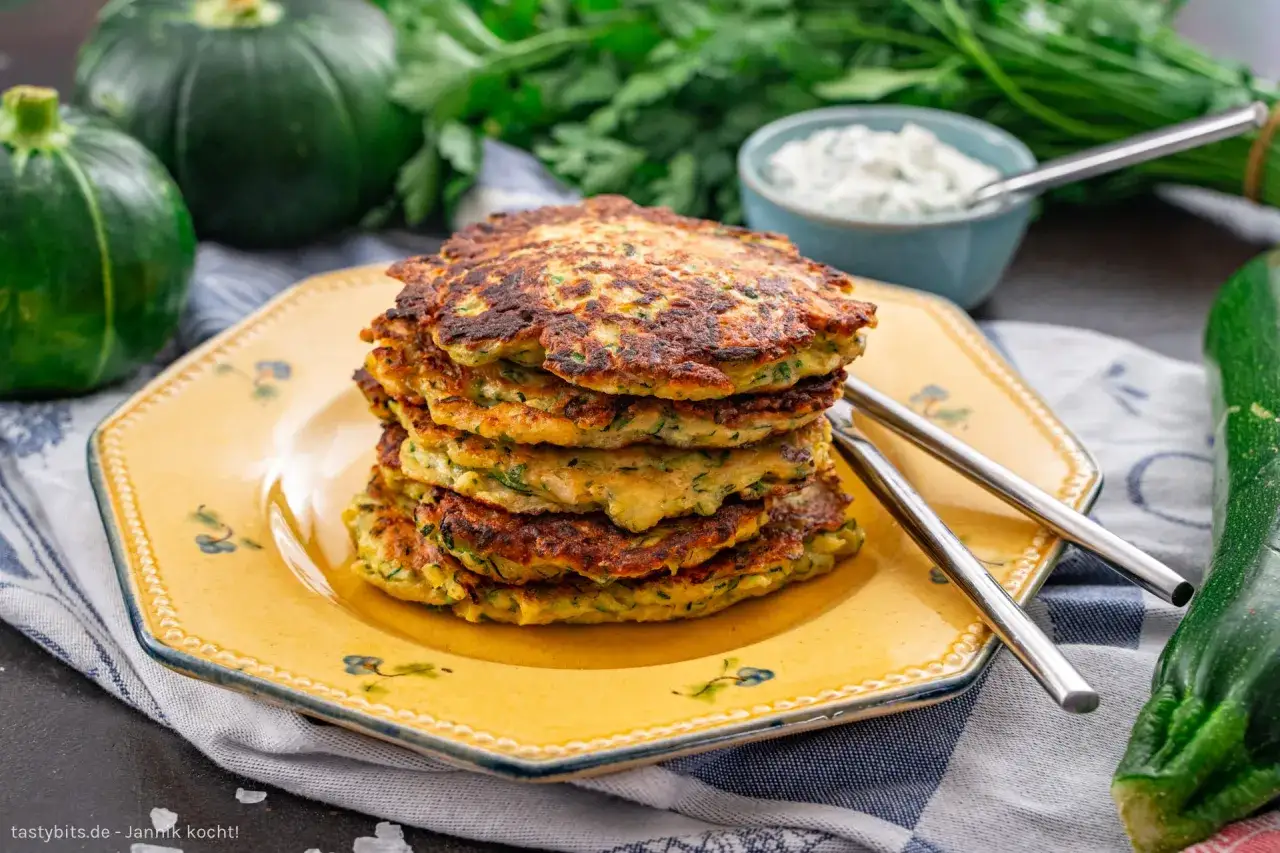 Stapel goldbrauner Zucchini-Puffer auf gelbem Teller, daneben frische Zucchini und Kr&auml;uter. Dazu ein Sch&auml;lchen Dip.