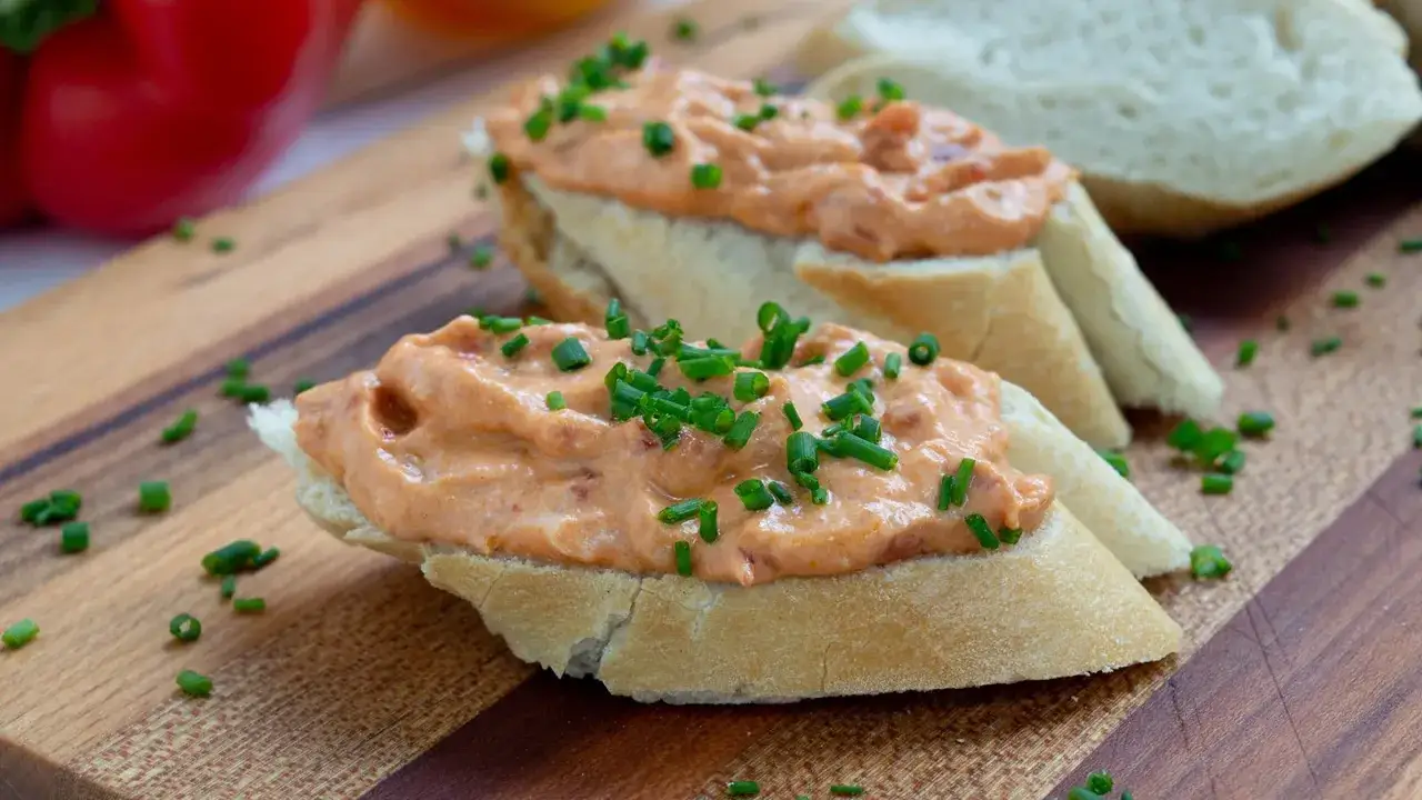 Baguettescheiben mit cremigem Ajvar Feta Dip und Schnittlauch bestreut, auf einem Holzbrett.