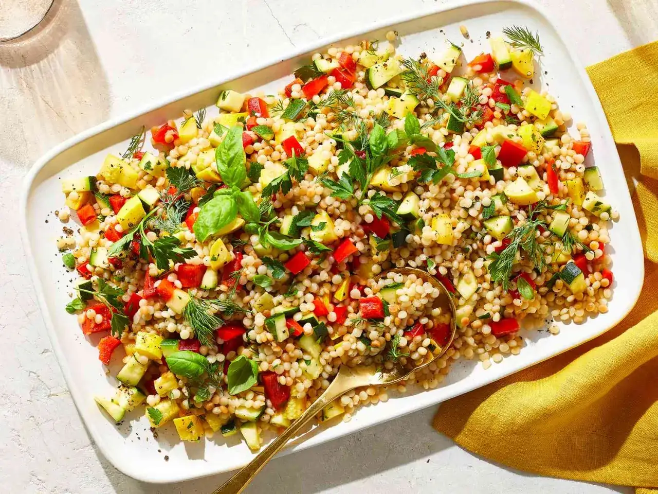 Bunter Couscous-Salat mit Paprika, Zucchini und Kräutern auf einer weißen Platte, garniert mit frischem Basilikum.
