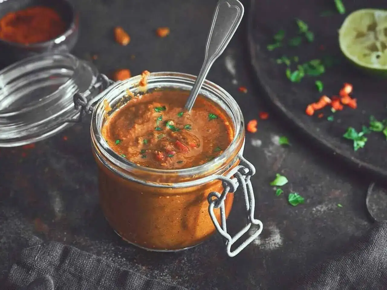 Ein Glas mit w&uuml;rziger Piri Piri Sauce, garniert mit frischen Kr&auml;utern. Daneben liegen Chilist&uuml;cke und eine Limettenspalte.