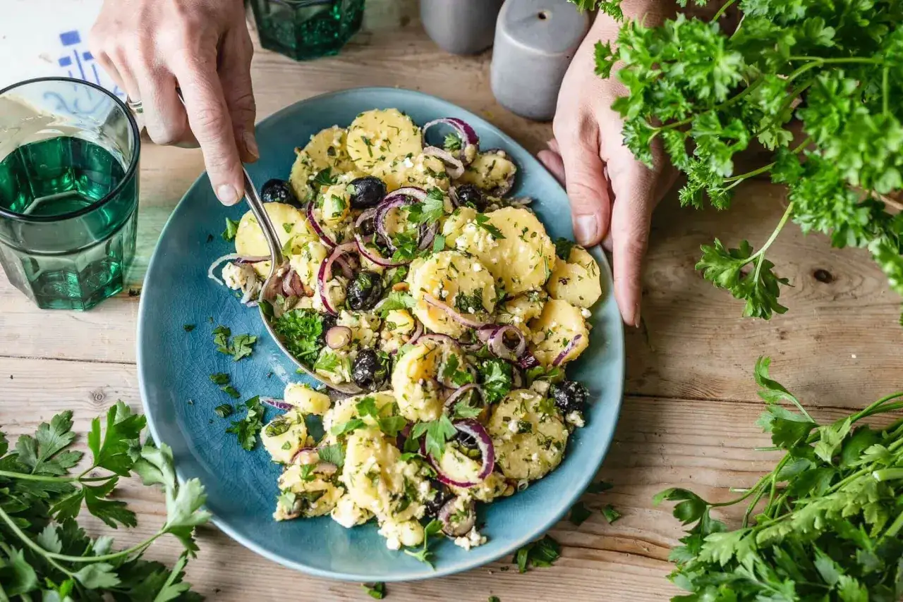 Kartoffelsalat mal anders: Kartoffelscheiben mit Oliven, roten Zwiebeln, Feta und frischer Petersilie auf blauem Teller.