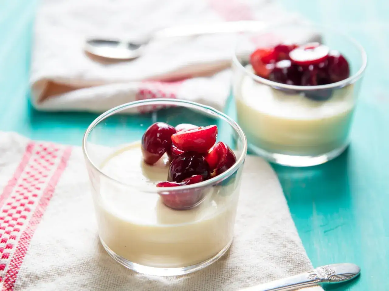 Zwei Gl&auml;ser cremiger Panna cotta, garniert mit frischen Kirschen, stehen auf einem rustikalen Holztisch.