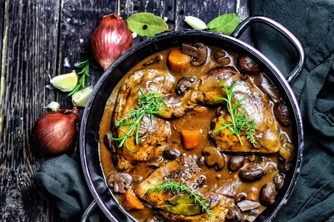 Herzhaftes Coq au Vin in einer Pfanne, mit Pilzen, Karotten und Kr&auml;utern garniert. Daneben Zwiebeln und Knoblauch.