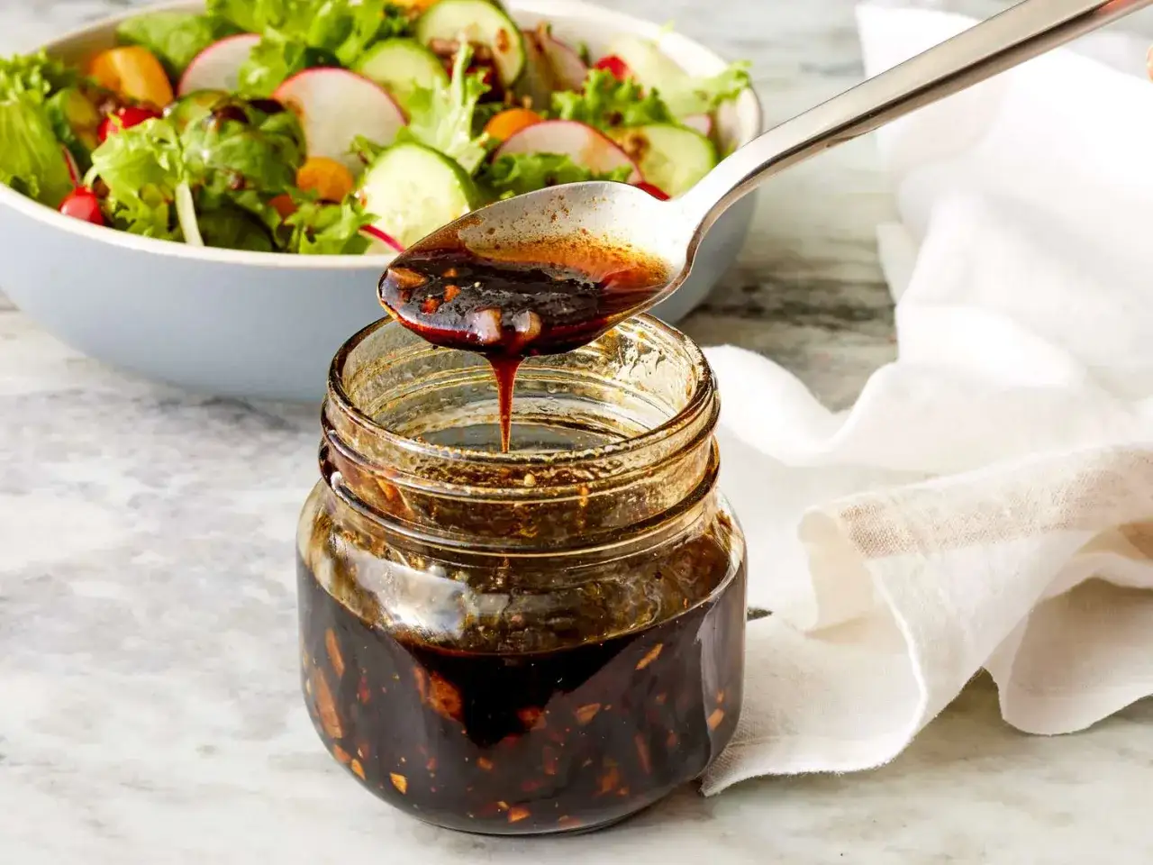 Ein L&ouml;ffel mit dunklem, gl&auml;nzendem Balsamico-Dressing tropft in ein Glas. Im Hintergrund ein frischer Salat.