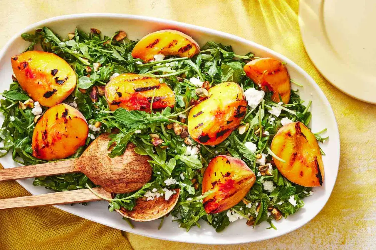Ein erfrischender Pfirsich Feta Salat mit Rucola, Pistazien und Minze, serviert auf einer ovalen Schale mit Holzservierl&ouml;ffeln.