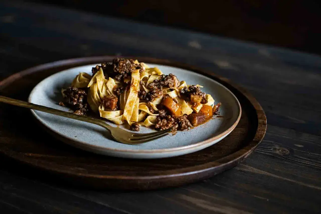 Ein Teller mit breiten Bandnudeln, bedeckt mit einer herzhaften Bolognese-Sauce und einigen K&uuml;rbisst&uuml;cken. Eine goldene Gabel liegt daneben.