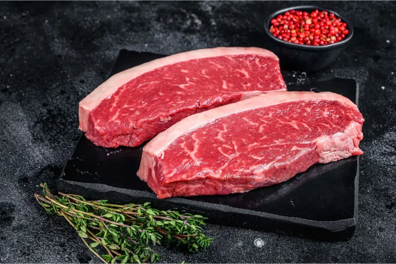 Zwei rohe Rindersteaks liegen auf einem schwarzen Stein. Perfekt, um sie medium rare zu braten. Daneben ein Zweig Thymian und eine Schale mit rosa Pfefferk&ouml;rnern.