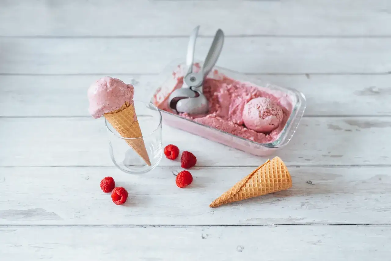 Himbeer Joghurt Eis in einer Waffel, daneben frische Himbeeren und eine Schale mit Eis.
