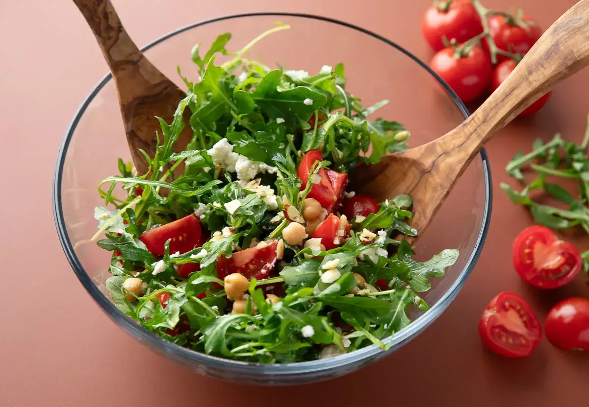 Frischer Rucolasalat mit Tomaten, Kichererbsen und Feta, serviert in einer Glassch&uuml;ssel mit Holzschaufeln.