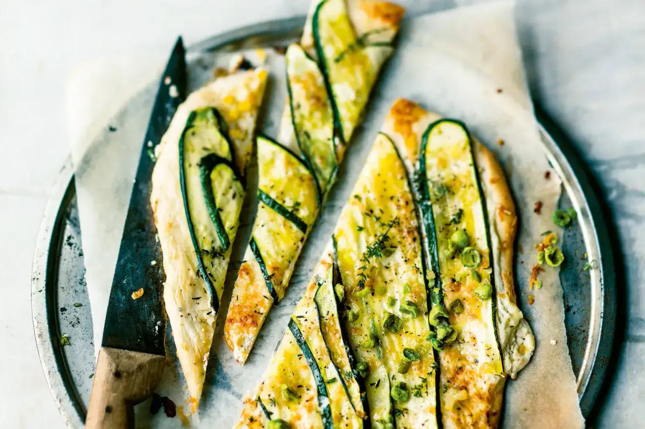 D&uuml;nner Flammkuchen mit Zucchini, K&auml;se und Kr&auml;utern, in St&uuml;cke geschnitten, auf einem Blech mit einem Messer.
