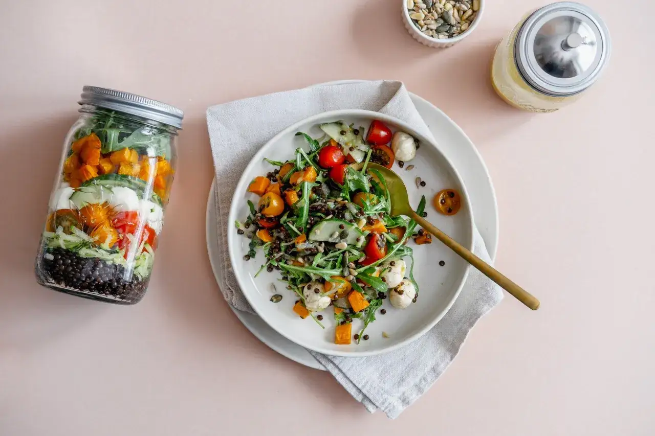 Schichtsalat im Glas mit Linsen, K&uuml;rbis, Tomaten und Mozzarella. Daneben ein Teller mit dem fertigen Salat und ein Glas Dressing.